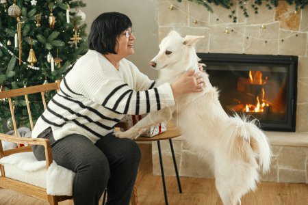 Téléchargez les photos : Belle femme mature jouant avec chien blanc mignon sur fond d'arbre de Noël élégant et cheminée festive. Heureuse femme adulte profitant de vacances d'hiver confortables avec animal de compagnie - en image libre de droit