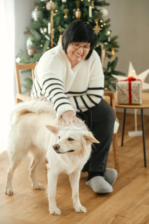 Foto de Feliz mujer mayor jugando con adorable perro blanco en la elegante sala de estar navideña. Hermosa mujer disfrutando de acogedoras vacaciones de invierno con linda mascota en el fondo del árbol de Navidad - Imagen libre de derechos