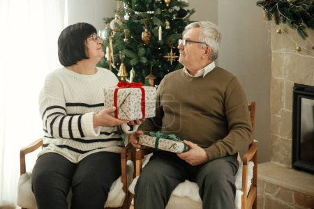 Téléchargez les photos : Beau couple de personnes âgées échangeant des cadeaux de Noël et assis à la cheminée chaude dans le salon décoré élégant et festif. Famille aînée avec une émotion sérieuse drôle profiter d'une soirée confortable avec des cadeaux - en image libre de droit