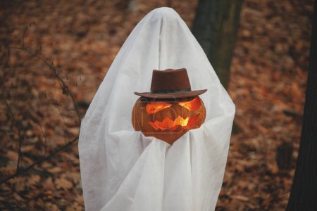 Téléchargez les photos : Joyeux Halloween ! Élégant fantôme tenant Jack o lanterne dans la forêt d'automne humide. Personne habillée en drap blanc comme fantôme avec citrouille debout dans les bois d'automne du soir. Trick ou traiter - en image libre de droit