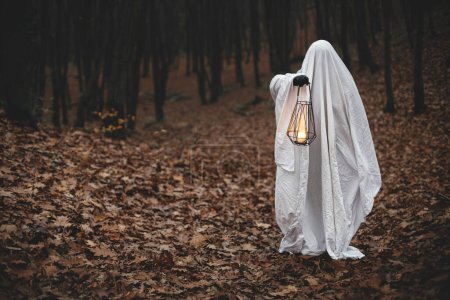 Téléchargez les photos : Fantôme effrayant tenant lanterne lumineuse dans la forêt d'automne sombre et humide. Personne habillée en drap blanc comme fantôme avec de la lumière dans les bois d'automne du soir. Joyeux Halloween ! Trick ou traiter - en image libre de droit
