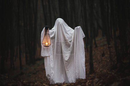 Téléchargez les photos : Joyeux Halloween ! Fantôme effrayant tenant lanterne lumineuse dans la forêt d'automne sombre et humide. Personne habillée en drap blanc comme fantôme avec de la lumière dans les bois d'automne du soir. Boum ! Temps d'horreur - en image libre de droit