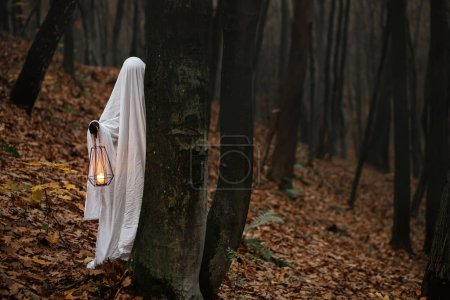 Photo for Happy Halloween! Spooky ghost holding glowing lantern and peeking out of a tree in moody dark autumn forest. Person dressed in white sheet as ghost in evening fall woods. Boo! Horror time - Royalty Free Image