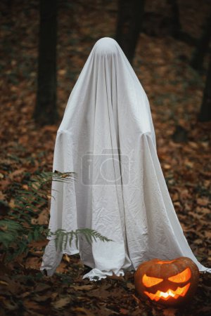 Téléchargez les photos : Joyeux Halloween ! Fantôme effrayant avec Jack o lanterne rougeoyante dans la forêt d'automne sombre et humide. Personne habillée en drap blanc comme fantôme avec citrouille debout dans les bois d'automne du soir. Trick ou traiter - en image libre de droit