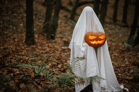 Téléchargez les photos : Fantôme effrayant tenant Jack o lanterne rougeoyante dans la forêt d'automne sombre et humide. Personne habillée en drap blanc comme fantôme avec citrouille debout dans les bois d'automne du soir. Joyeux Halloween ! Trick ou traiter - en image libre de droit
