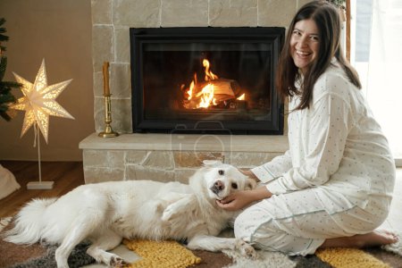 Téléchargez les photos : Belle femme en pyjama élégant relaxant avec chien blanc mignon à la cheminée confortable, profitant du matin de Noël calme dans le salon décoré festif. Joyeux Noël ! Vacances d'hiver avec animaux domestiques - en image libre de droit