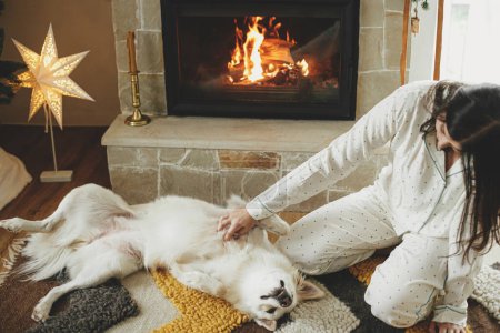 Foto de Acogedora mañana de Navidad. Hermosa mujer en pijama elegante relajarse y jugar con lindo perro blanco en la chimenea caliente en la sala de estar decorada festiva. ¡Feliz Navidad! Vacaciones de invierno con mascota - Imagen libre de derechos