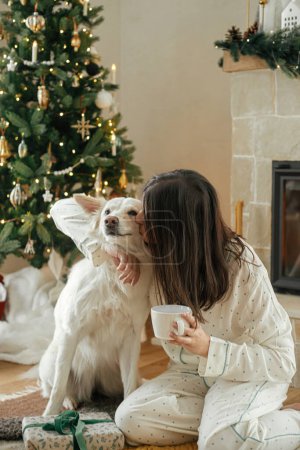 Téléchargez les photos : Confortable matin de Noël. Belle femme en pyjama tenant du thé chaud et étreignant chien mignon à la cheminée dans le salon décoré festif. Joyeux Noël ! Vacances d'hiver avec animaux domestiques - en image libre de droit