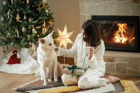 Photo for Cozy christmas morning. Beautiful woman in stylish pajamas holding warm tea and relaxing with cute  dog at fireplace in festive decorated living room. Merry Christmas! Winter holidays with pet - Royalty Free Image