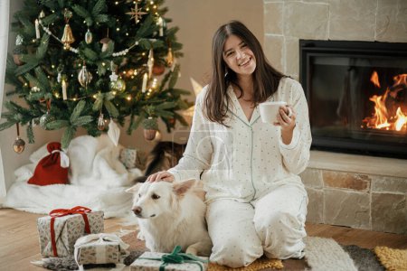Téléchargez les photos : Belle femme en pyjama élégant avec thé chaud relaxant avec chien mignon à la cheminée confortable, profiter du matin de Noël dans le salon décoré de fêtes avec des cadeaux. Joyeux Noël ! Vacances d'hiver - en image libre de droit