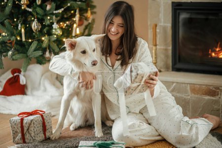 Photo for Beautiful woman in pajamas holding stylish christmas gift and hugging cute white dog at decorated tree and fireplace, enjoying cozy christmas morning in festive living room. Merry Christmas - Royalty Free Image