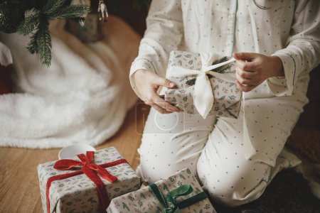 Téléchargez les photos : Femme élégante en pyjama ouvrant cadeau de Noël enveloppé moderne sous arbre décoré fermer, profiter matin de Noël confortable dans le salon festif. Joyeux Noël - en image libre de droit