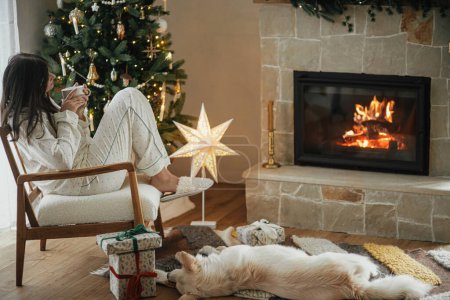 Téléchargez les photos : Confortable matin de Noël. Belle femme en pyjama élégant relaxant avec thé chaud et chien mignon à la cheminée dans le salon décoré festif. Joyeux Noël ! Vacances d'hiver avec animaux domestiques - en image libre de droit