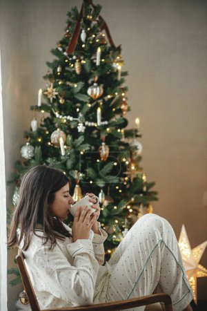 Téléchargez les photos : Belle femme en pyjama élégant avec thé chaud relaxant à l'arbre décoré, profiter du matin de Noël confortable dans le salon de fête. Joyeux Noël ! Hygge hivernale - en image libre de droit