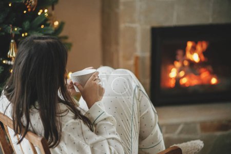 Foto de Hermosa mujer en pijama con estilo con té caliente relajante en el árbol decorado y la chimenea, disfrutando de una acogedora mañana de Navidad en la sala de estar festiva. ¡Feliz Navidad! Hygge de invierno - Imagen libre de derechos