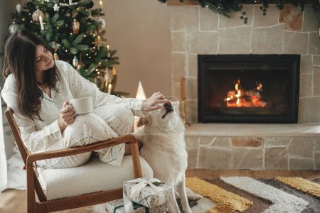 Téléchargez les photos : Confortable matin de Noël. Belle femme en pyjama élégant relaxant avec thé chaud et chien mignon à la cheminée dans le salon décoré festif. Joyeux Noël ! Vacances d'hiver avec animaux domestiques - en image libre de droit
