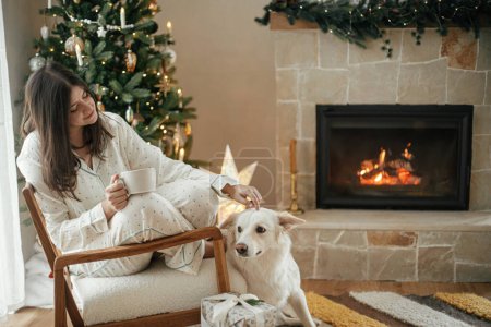 Foto de Hermosa mujer en pijama con estilo con té caliente relajante en la acogedora chimenea con perro lindo, disfrutando de la mañana de Navidad en la sala de estar decorada festiva con regalos. ¡Feliz Navidad! Hygge de invierno - Imagen libre de derechos