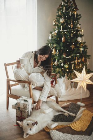 Foto de Hermosa mujer en pijama con estilo con té caliente relajante en el árbol moderno con perro lindo, disfrutando de la mañana de Navidad en la sala de estar festiva con regalos. ¡Feliz Navidad! Hygge de invierno - Imagen libre de derechos