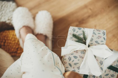 Photo for Cozy Christmas morning. Stylish christmas gift with ribbon and fir branch and woman legs in cozy slippers top view in festive living room. Merry Christmas and happy holidays! - Royalty Free Image