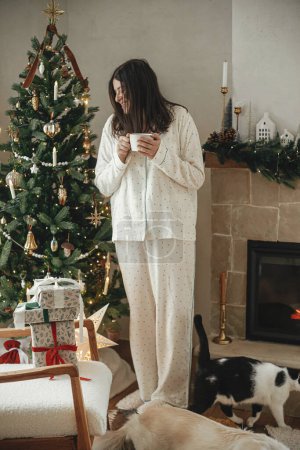 Téléchargez les photos : Confortable matin de Noël. Belle femme en pyjama élégant relaxant avec thé chaud et animaux mignons à la cheminée dans le salon décoré festive. Joyeux Noël ! Vacances d'hiver avec animaux domestiques - en image libre de droit