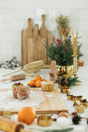 Foto de Masa de pan de jengibre con harina sobre tabla de madera, rodillo, cortadores de metal dorado, especias de cocina y decoraciones festivas en la encimera de la cocina blanca moderna. Hacer galletas de jengibre de Navidad - Imagen libre de derechos
