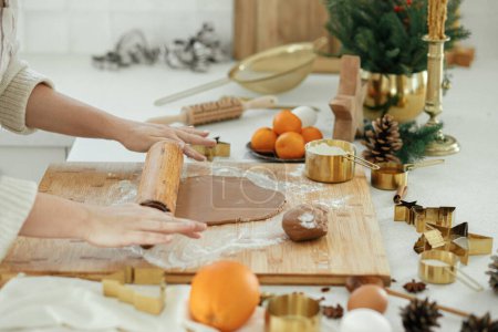 Téléchargez les photos : Mains pétrissant la pâte de pain d'épice sur une planche en bois avec rouleau à pâtisserie, coupe-métal doré, épices de cuisine, décorations festives dans la cuisine blanche moderne. Femme faisant des biscuits de pain d'épice de Noël - en image libre de droit