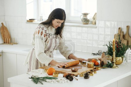 Foto de Mujer haciendo galletas de jengibre de Navidad en la cocina blanca moderna. Mano amasando masa de jengibre en tablero de madera con harina, rodillo, cortadores de metal dorado, especias de cocina en la encimera - Imagen libre de derechos