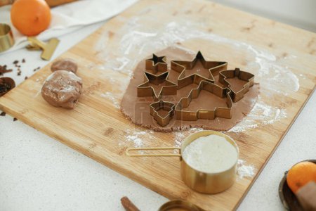 Téléchargez les photos : Pâte à pain d'épice avec découpeuses métalliques dorées festives sur planche de bois avec farine, épices de cuisson, décorations festives dans la cuisine blanche moderne. Faire des biscuits au pain d'épice de Noël - en image libre de droit