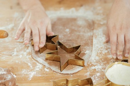Foto de Manos cortando masa de pan de jengibre con cortadores de metal dorado festivos en tablero de madera con harina, especias de cocina en la encimera en la cocina blanca moderna. Mujer haciendo galletas de jengibre de Navidad - Imagen libre de derechos