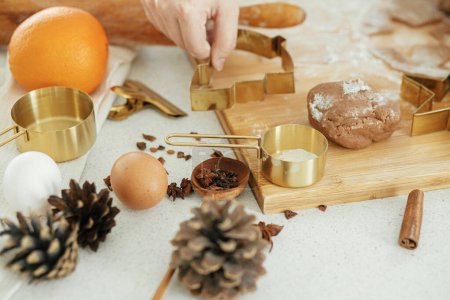 Foto de Hacer galletas de jengibre de Navidad. Masa de pan de jengibre con cortadores de metal dorado festivos en tablero de madera con harina, especias de cocina, decoraciones festivas en la cocina blanca moderna - Imagen libre de derechos