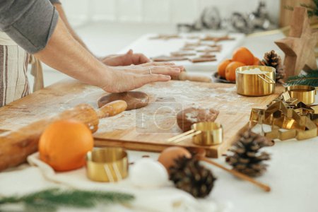 Foto de Hombre haciendo galletas de jengibre de Navidad en la moderna cocina blanca de cerca. Manos amasando masa de pan de jengibre con rodillo, cortadores de metal dorado, especias de cocina, decoraciones festivas - Imagen libre de derechos