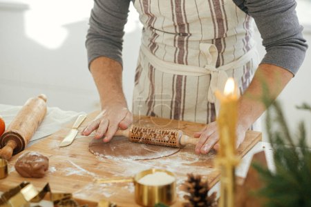Foto de Hombre haciendo galletas de jengibre de Navidad en la cocina blanca moderna. Mano amasando masa de jengibre en tablero de madera con harina, rodillo, cortadores de metal dorado, especias de cocina en la encimera - Imagen libre de derechos