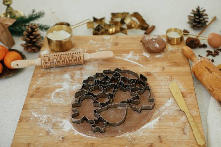 Photo for Making christmas gingerbread cookies. Gingerbread dough with festive metal cutters on wooden board with flour, cooking spices, festive decorations in modern white kitchen - Royalty Free Image
