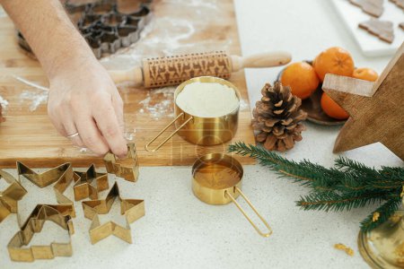 Foto de Manos cortando masa de pan de jengibre con cortadores de metal dorado festivos en tablero de madera con harina, especias de cocina en la encimera en la cocina blanca moderna. Hombre haciendo galletas de jengibre de Navidad - Imagen libre de derechos