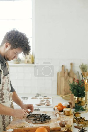 Téléchargez les photos : Homme faisant des biscuits de pain d'épice de Noël fermer dans la cuisine blanche moderne. Mains coupant la pâte de pain d'épice avec des coupeurs de métal festifs avec des épices et des décorations de cuisson - en image libre de droit