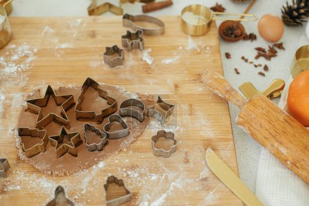 Foto de Masa de pan de jengibre con cortadores de metal dorado festivos en tablero de madera con harina, especias de cocina, decoraciones festivas en la cocina blanca moderna. Hacer galletas de jengibre de Navidad - Imagen libre de derechos