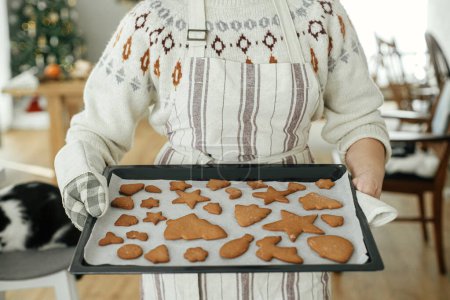 Foto de Mujer en delantal y manoplas de horno sosteniendo bandeja con galletas de jengibre recién horneadas en formas festivas de Navidad en la moderna cocina blanca. Hacer galletas de jengibre de Navidad - Imagen libre de derechos