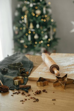 Téléchargez les photos : Je fais des biscuits au pain d'épice de Noël. Farine sur panneau de bois, rouleau à pâtisserie, coupe-métal doré, épices de cuisine et décorations festives sur table rustique contre l'arbre de Noël décoré élégant - en image libre de droit