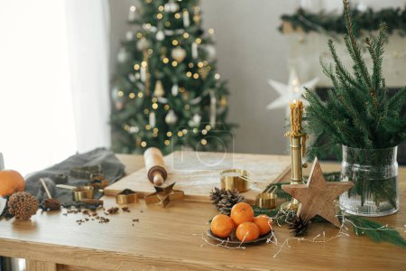 Foto de Hacer galletas de jengibre de Navidad. Estrella de madera, naranjas, cortadores de metal dorado, especias de cocina y decoraciones festivas en la mesa rústica contra el elegante árbol de Navidad decorado - Imagen libre de derechos