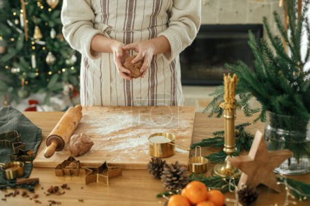 Foto de Mujer haciendo galletas de jengibre de Navidad. Manos amasando masa de pan de jengibre con rodillo, cortadores de oro, especias de cocina, decoraciones festivas en la mesa rústica contra el elegante árbol de Navidad - Imagen libre de derechos