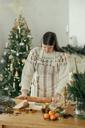 Téléchargez les photos : Mains pétrissant pâte à pain d'épice avec rouleau à pâtisserie, épices de cuisson, décorations festives sur table rustique contre l'arbre de Noël élégant. Femme faisant des biscuits de pain d'épice de Noël, temps des fêtes - en image libre de droit