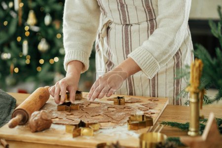 Téléchargez les photos : Femme faisant des biscuits de pain d'épice de Noël, l'heure de l'atmosphère. Mains coupant la pâte de pain d'épice avec des coupeuses dorées festives sur la table rustique avec des décorations de vacances contre l'arbre de Noël élégant - en image libre de droit