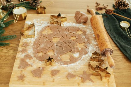 Photo for Making christmas gingerbread cookies, atmospheric holiday time. Gingerbread dough with golden cutters, rolling pin, fir branches and festive decorations on rustic wooden table - Royalty Free Image