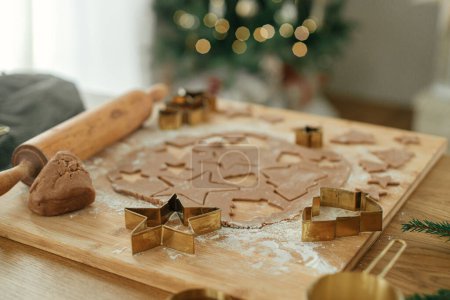 Téléchargez les photos : Je fais des biscuits au pain d'épice de Noël. Pâte à pain d'épice avec couteaux dorés sur planche en bois, rouleau à pâtisserie, épices de cuisson et décorations festives sur table rustique contre l'arbre de Noël élégant - en image libre de droit
