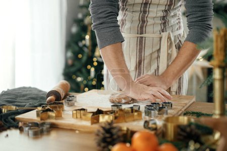 Téléchargez les photos : Un homme qui fait des biscuits au pain d'épice de Noël. Mains pétrissant pâte à pain d'épice avec rouleau à pâtisserie, coupeuses dorées, épices de cuisson, décorations festives sur table rustique contre l'arbre de Noël élégant - en image libre de droit