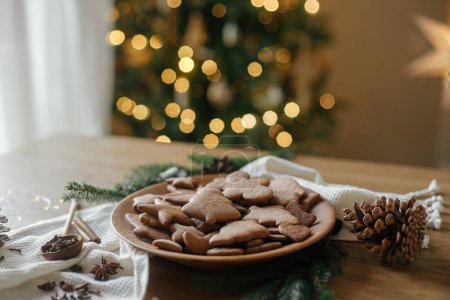 Téléchargez les photos : Biscuits de pain d'épice de Noël dans une assiette en bois sur une table rustique sur fond d'éclairage élégant de Noël doré. Joyeux Noël ! Délicieux biscuits au pain d'épice, vacances atmosphériques - en image libre de droit