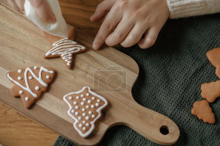 Téléchargez les photos : Décorer les biscuits au pain d'épice avec la cerise sur la table en bois rustique fermer. Traditions de Noël atmosphérique. Mains décorer les biscuits de Noël cuits au four avec du glaçage au sucre. Temps en famille - en image libre de droit