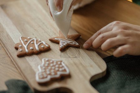 Photo for Hands decorating christmas gingerbread cookies with icing on rustic wooden table close up. Atmospheric Christmas holiday traditions. Decorating baked cookies with sugar frosting. Family time - Royalty Free Image