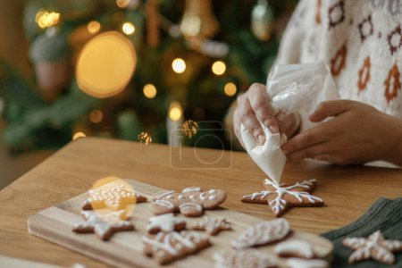 Foto de Manos decorando galletas de jengibre navideño con glaseado sobre mesa de madera rústica sobre fondo de luces doradas. Tradiciones navideñas atmosféricas. Decorar galletas con glaseado de azúcar - Imagen libre de derechos