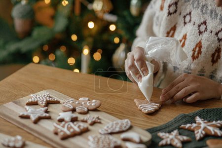 Téléchargez les photos : Mains décorer biscuits de pain d'épice de Noël avec cerise sur une table en bois rustique sur fond de lumières dorées. Traditions de Noël atmosphérique. Décorer les biscuits avec du glaçage au sucre - en image libre de droit